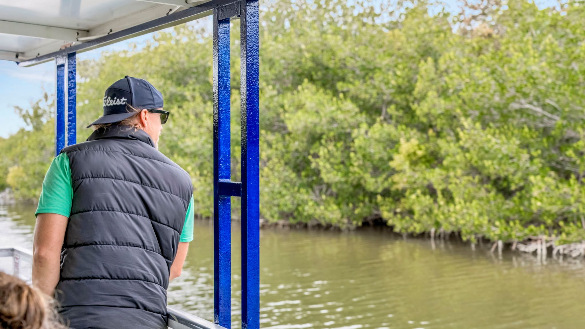 Person in a black vest and cap on a boat, looking at lush green mangroves.