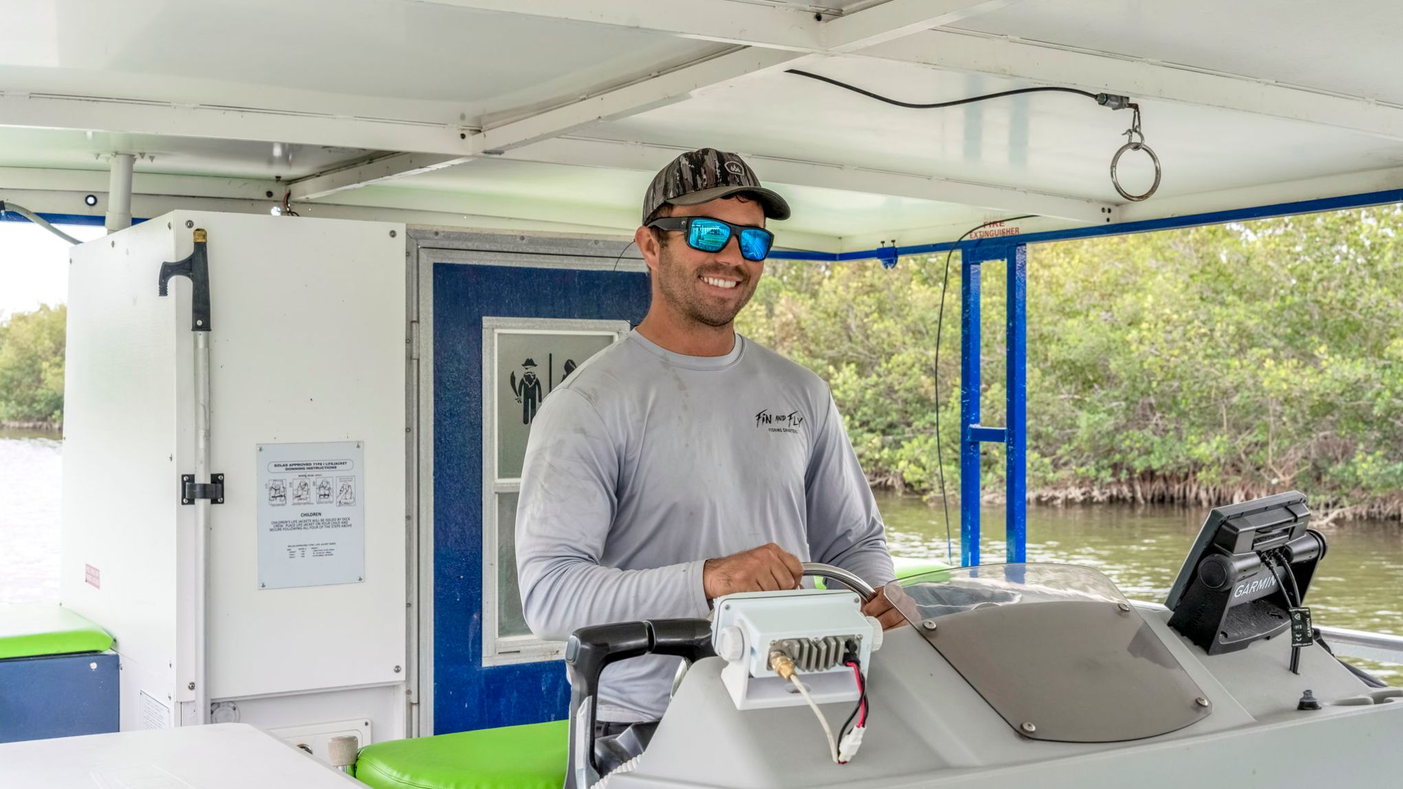 Man in sunglasses and hat steering a boat with green seats on a river.