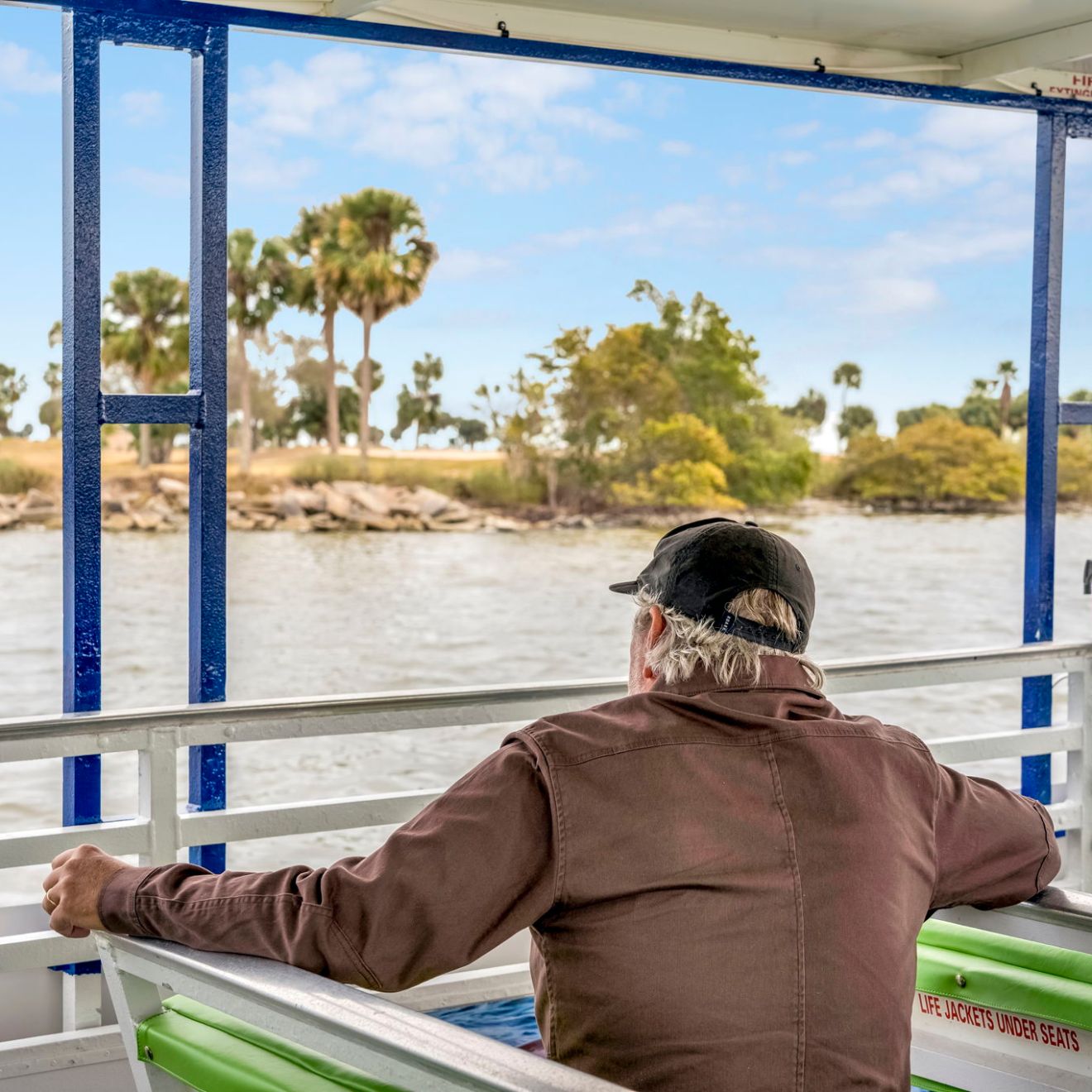 Man on a boat looking at a lake with palm trees under a blue sky.