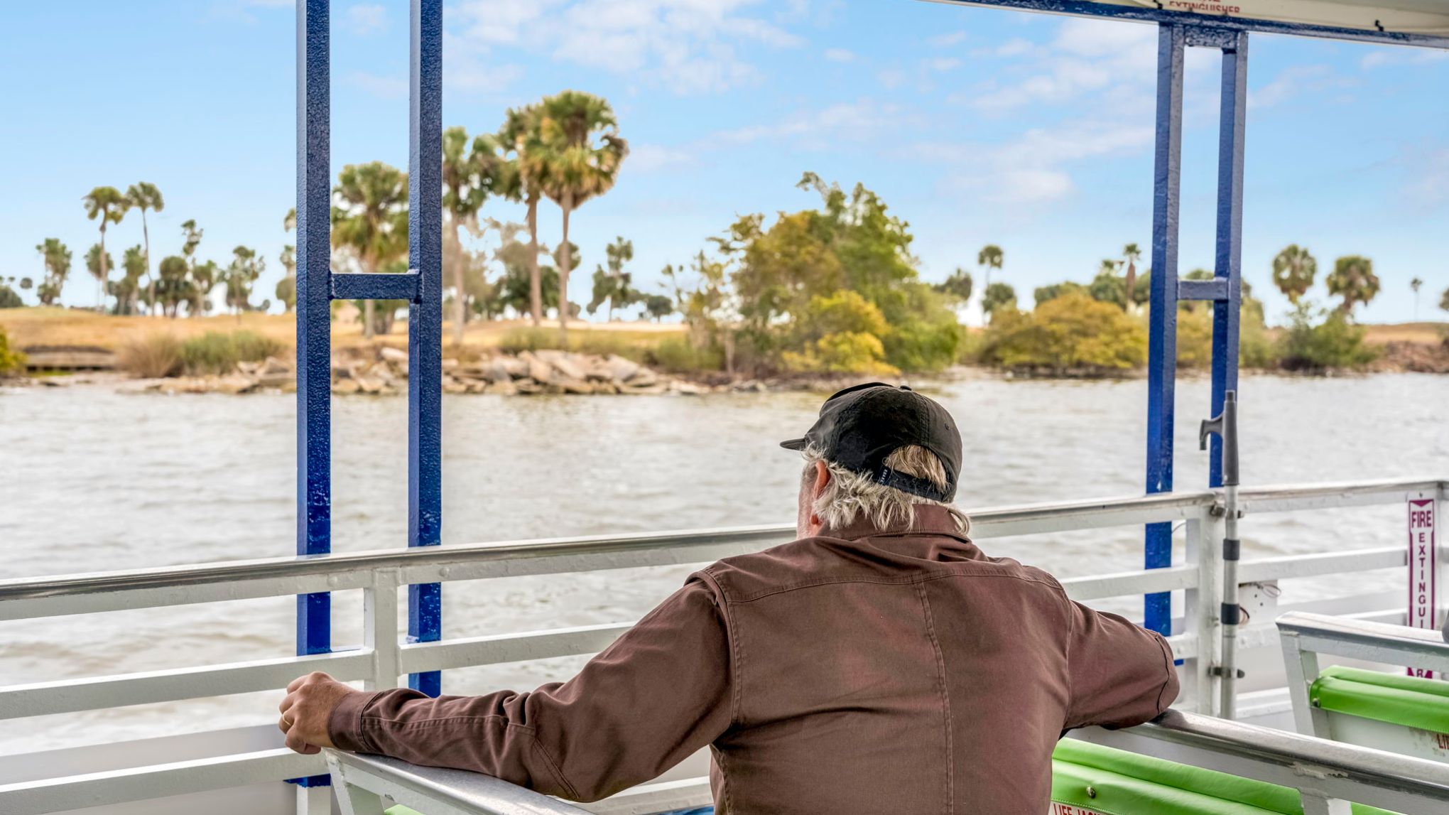 Man on a boat looking at a lake with palm trees under a blue sky.