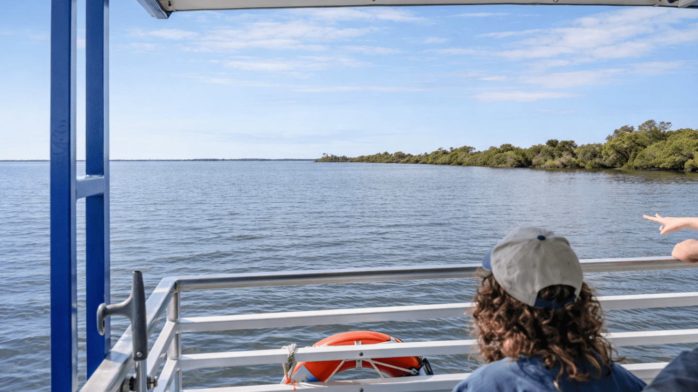 People on a boat looking at water and distant greenery under a partly cloudy sky.