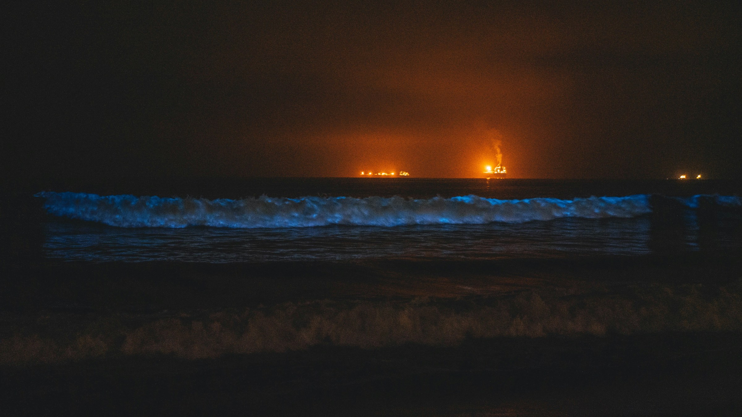 Bioluminescent waves glow blue at night, with distant oil rigs lit in orange on the horizon.